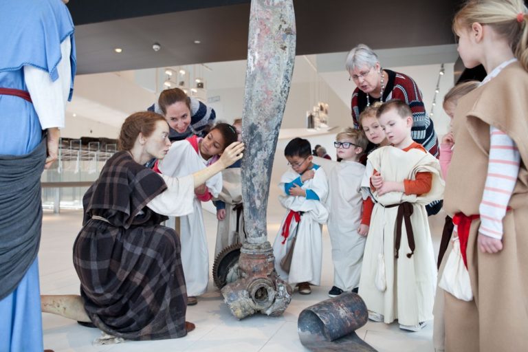 Een vrijwilliger staat met 8 kinderen in Romeinse kledij en een volwassene op zaal in Museum Hoge Woerd. Een omgekeerde blik in de tijd: Twee beelden van Romeinse vrouwen ontdekken een propeller.