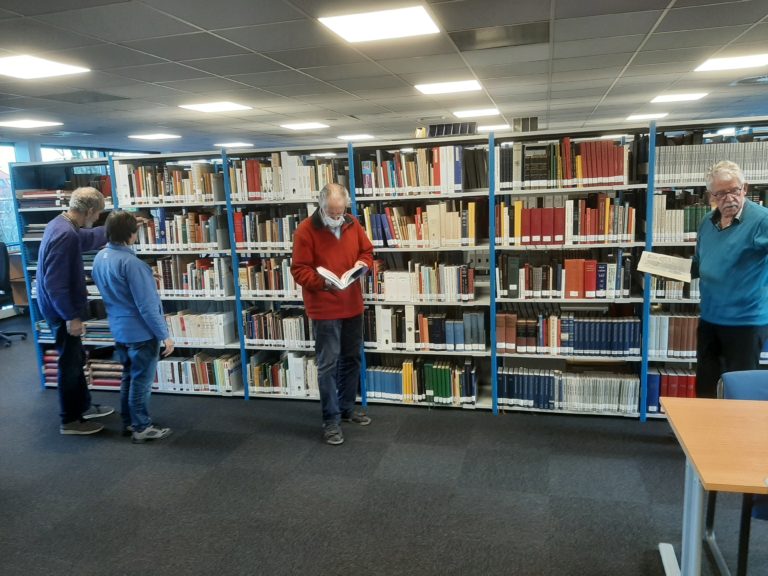 Vier mensen staan in een bibliotheek. Twee met een opengeslagen boek.