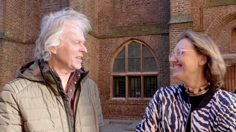 Erfgoedvrijwilligers Hans en Cecile kijken elkaar aan en staan voor een oud gebouw.