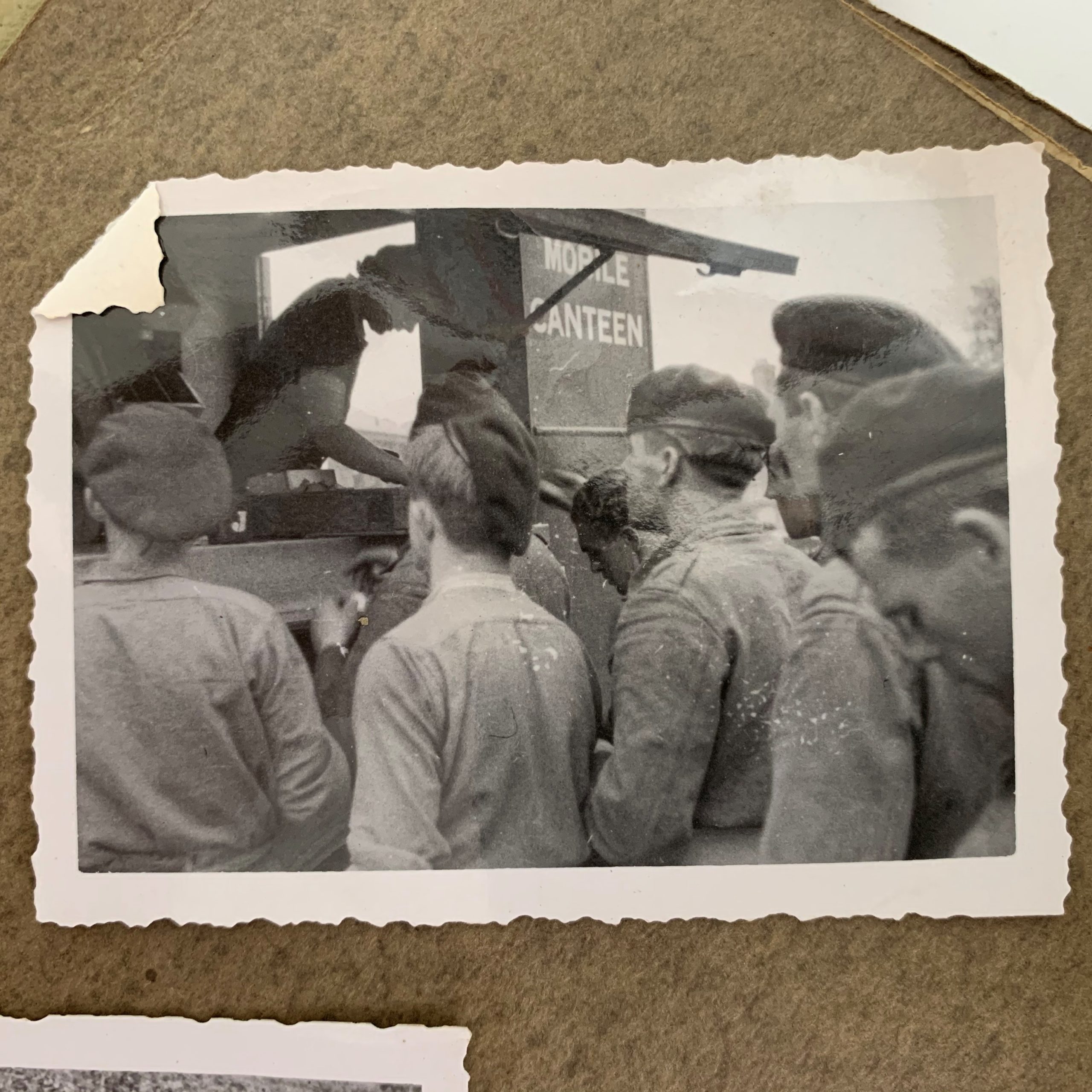 Een oude zwart-witte foto in een fotoboek geplakt waarop je de achterkant van soldaten met petten op ziet staan, ze staan gericht naar een kraam.