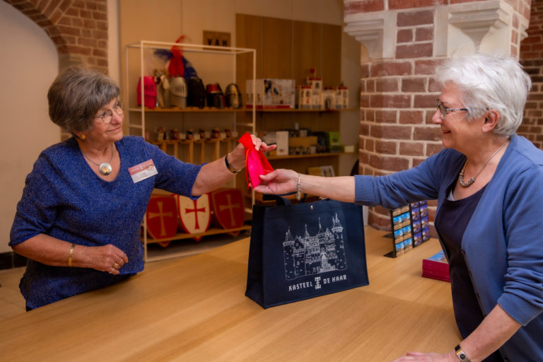 Een museumwinkel vrijwilligster overhandigd een ingepakt cadeautje aan een bezoeker.
