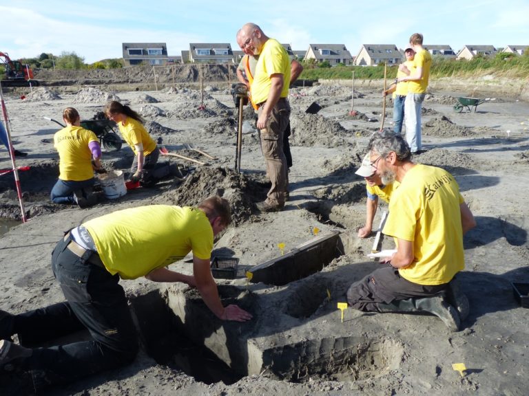 Sfeerbeeld van de opgraving Katwijk de Zanderij. De mensen op de foto hebben gele t-shirts aan en staan of zitten in een zandvlakte.
