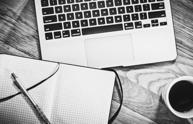 Zwart wit foto van een laptop met een kopje koffie en een leeg notitieboekje.