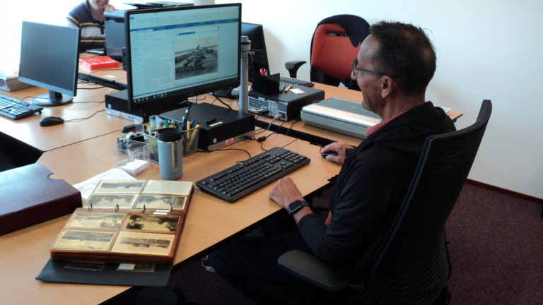 Man zit achter een computerscherm met op zijn bureau een map met oude foto's.