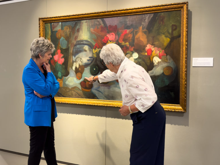 Een rondleidster en bezoeker staan samen bij een groot kleurrijk schilderij. De rondleidster wijst iets aan op het schilderij.