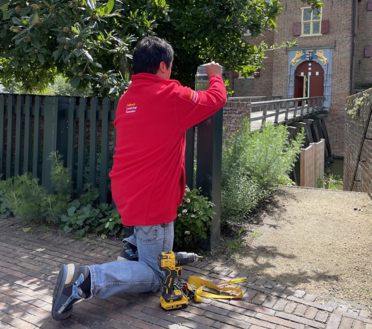 Vrijwilliger in rode jas klust met een boormachine aan het hek van het kasteel.