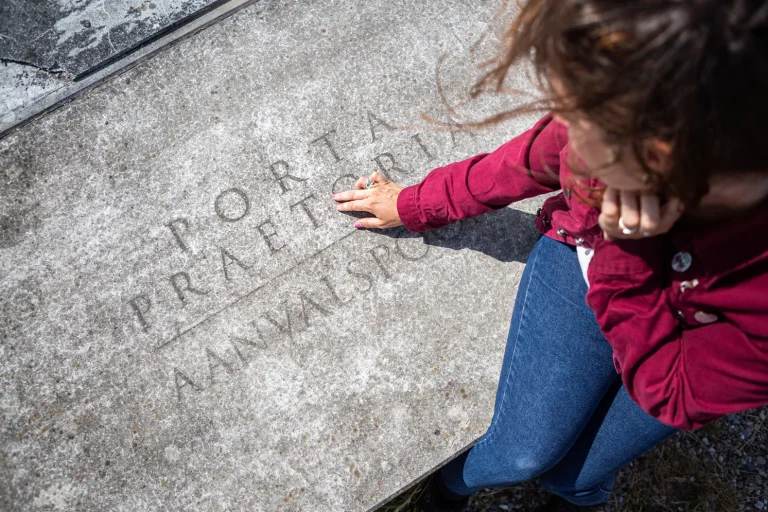 Een vrouw zit op een grijze platte grijze steen waar 'porta praetori' op uitgehouwen is.