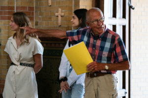 Een man wijst iets in de Sint Bonifatiuskerk en houdt een geel papier in zijn handen vast. Er staan twee bezoekers op de achtergrond.