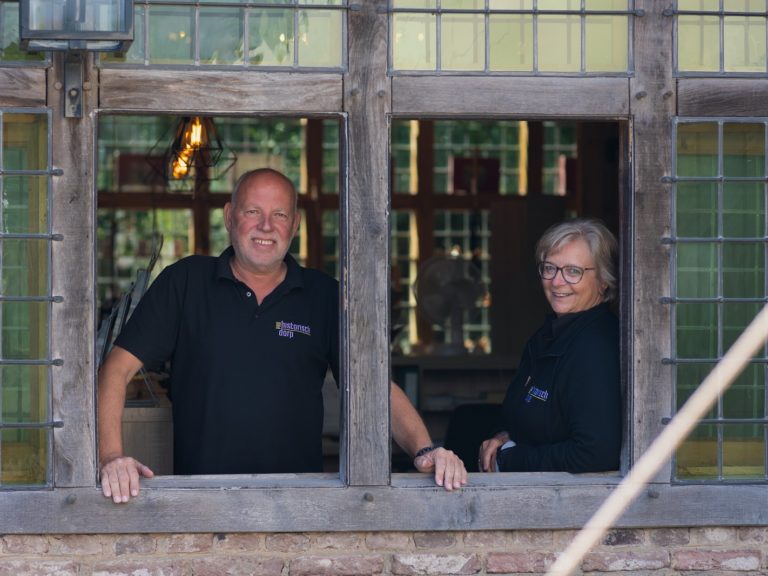 Twee mensen staan achter een oud raamkozijn en kijken lachend in de camera