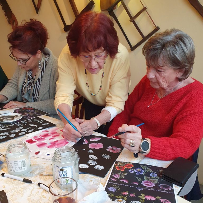 Drie oudere vrouwen zijn samen bezig met een workshop schilderen. Ze schilderen bloemen op zwart papier.