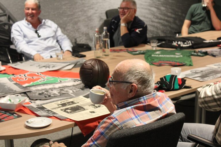 Oudere man zit aan tafel met enkele andere personen. Op de tafel liggen archiefstukken van het vroegere N.E.C. voetbal.