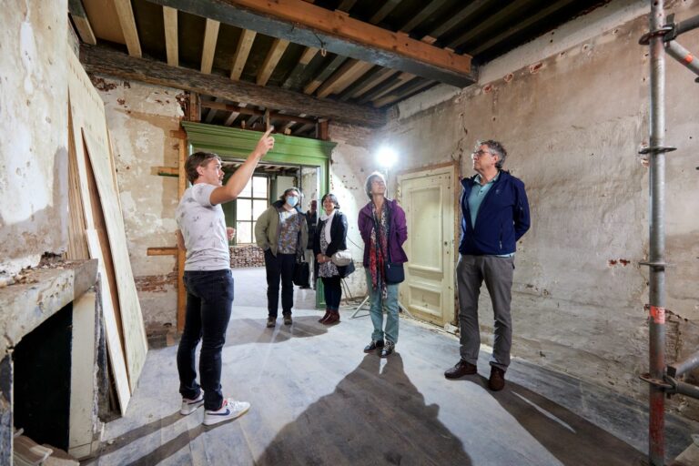 Een vrijwilliger geeft uitleg aan vier bezoekers in een monumentaal pand, waar restauratiewerkzaamheden plaatsvinden.